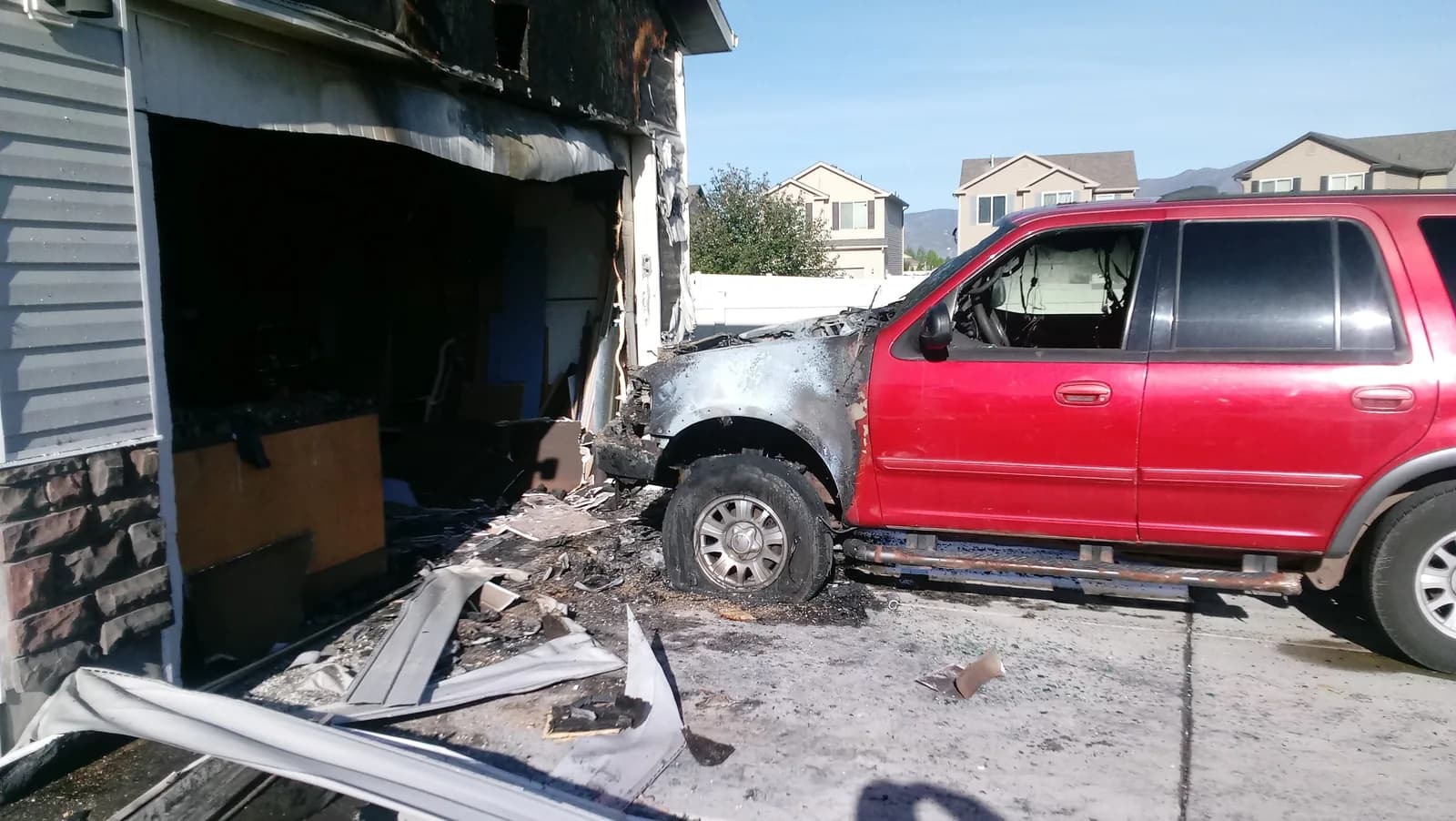 Fire-damaged garage with burned siding and debris beside the vehicle parked in the driveway